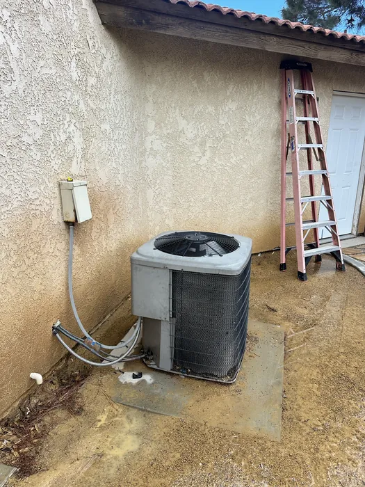 Wall-mounted AC condenser unit with refrigerant lineset on a stucco exterior wall beside an extension ladder at an Inland Empire home.