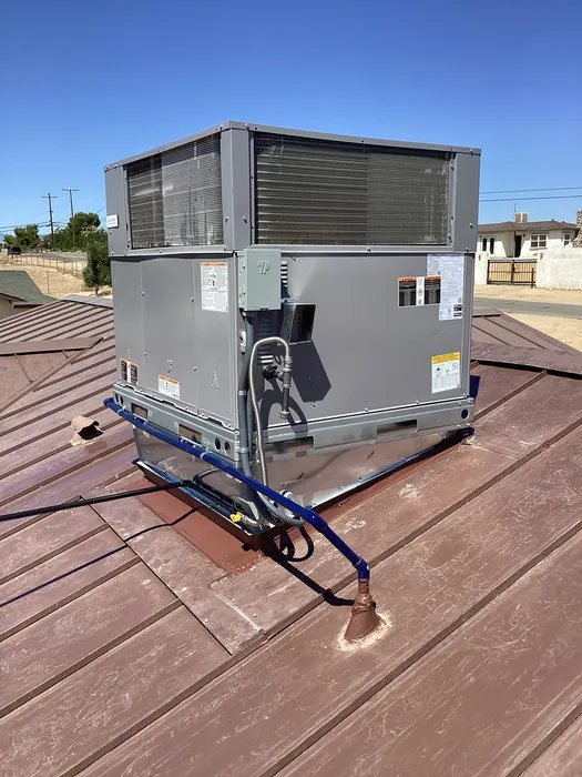 A gray packaged rooftop HVAC unit installed on a brown tile roof with a blue ladder hooked over the roof edge under a clear sky.