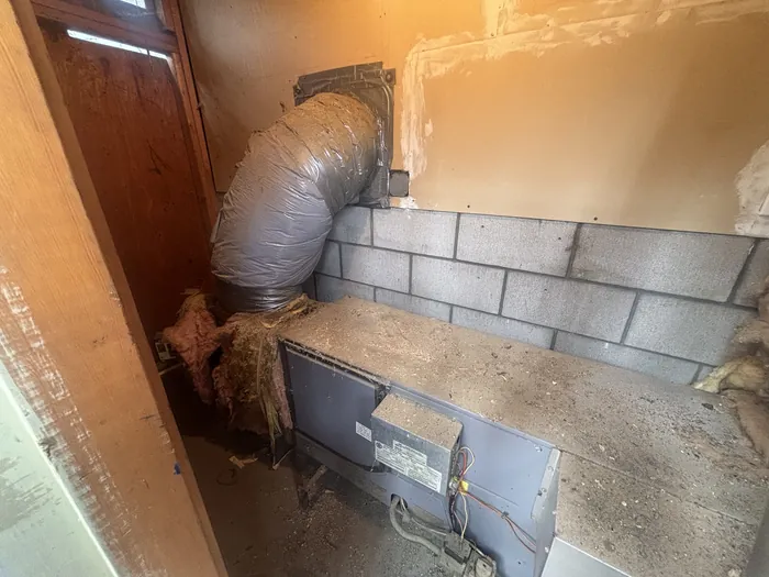 An aging horizontal gas furnace with a kinked gray flex duct connection sitting against a cinder block wall in a High Desert garage utility area.