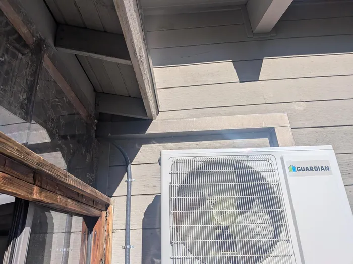 Guardian-brand mini-split condenser mounted on a gray board-and-batten exterior wall with line set running up under the eave at a High Desert home.